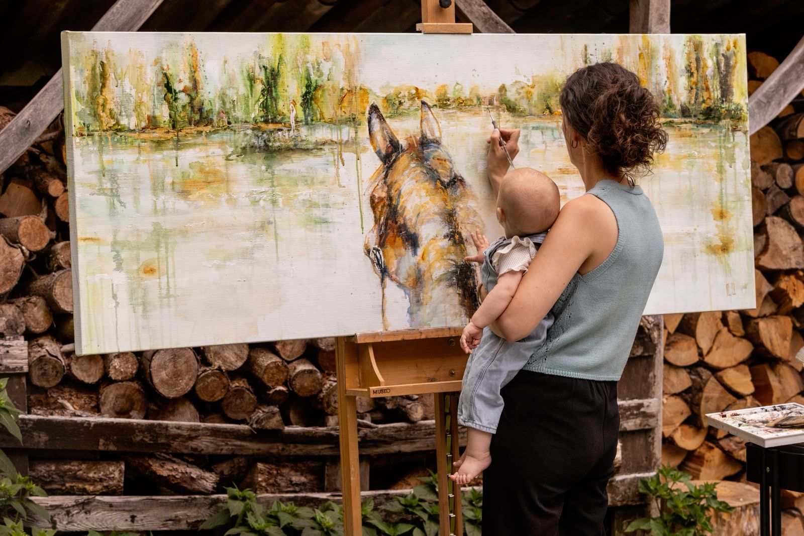 Marjo Lebbe painting a horse outdoors while holding her baby, who reaches toward the canvas
