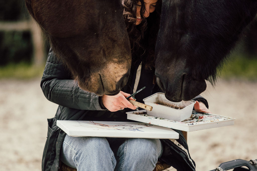Marjo painting with her palette while two horses lean in to her