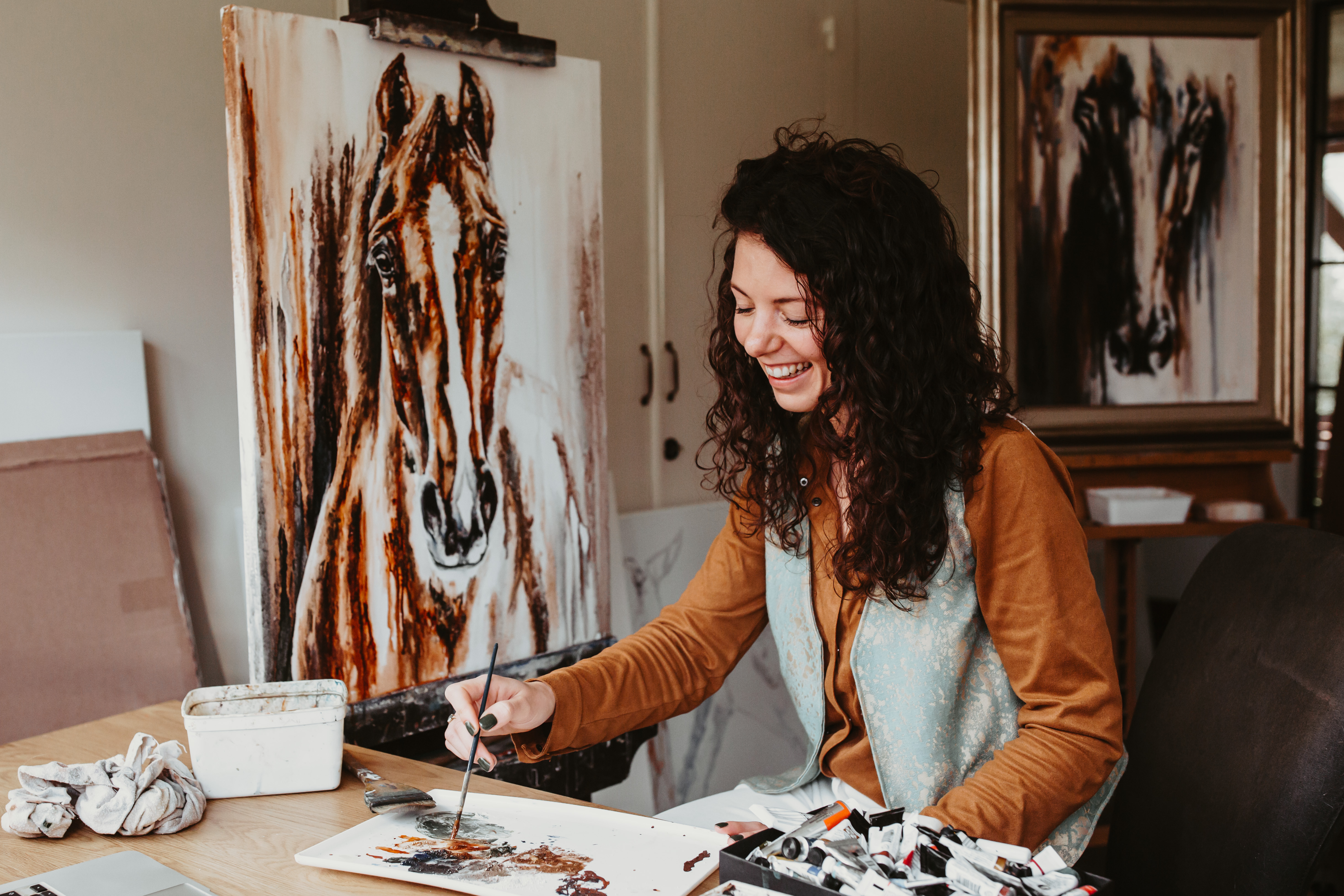 Marjo smiling as she mixes watercolour at her table, a horse portrait on the easel behind her