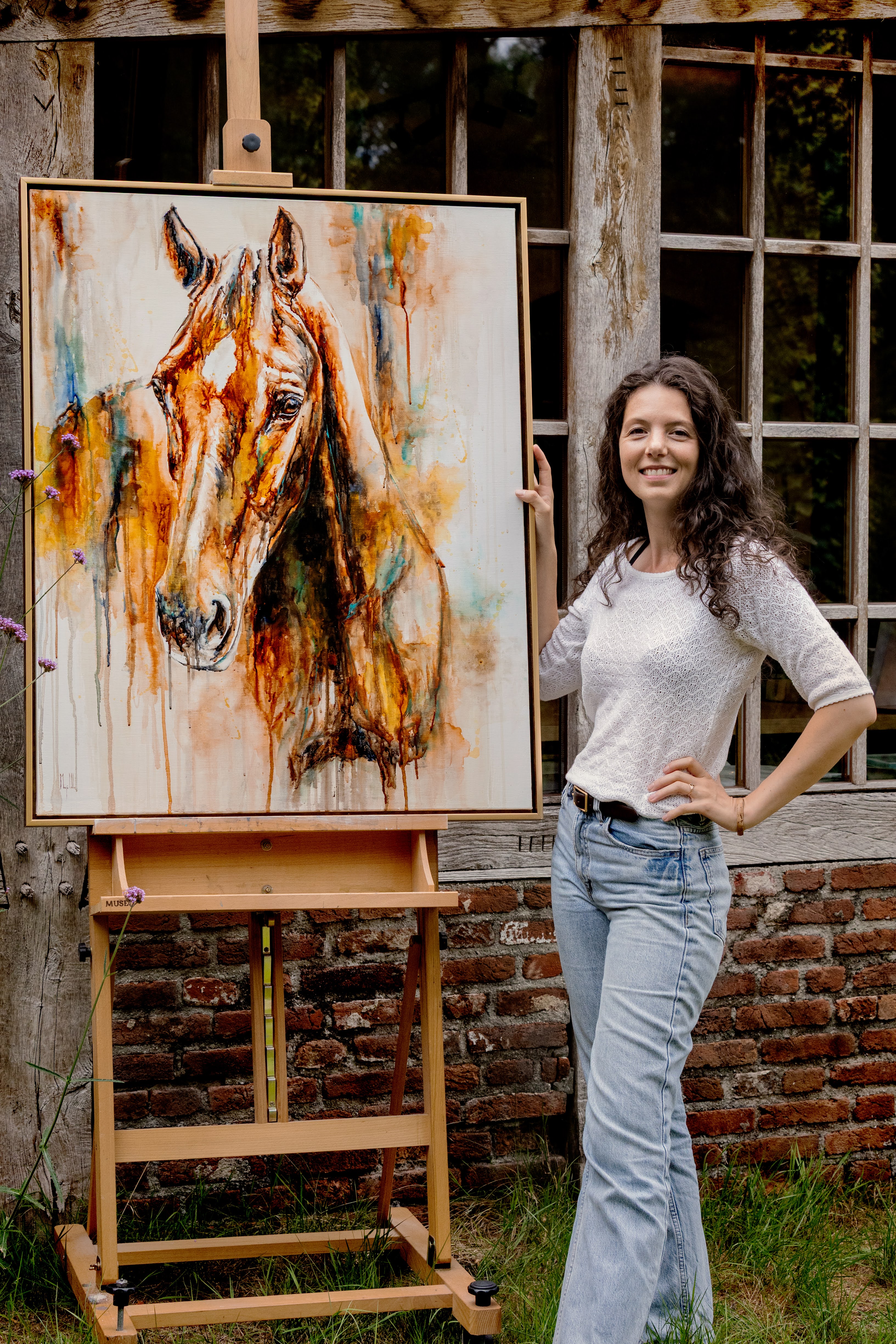 Marjo smiling beside an easel with a colourful horse portrait, outdoors against a rustic brick wall