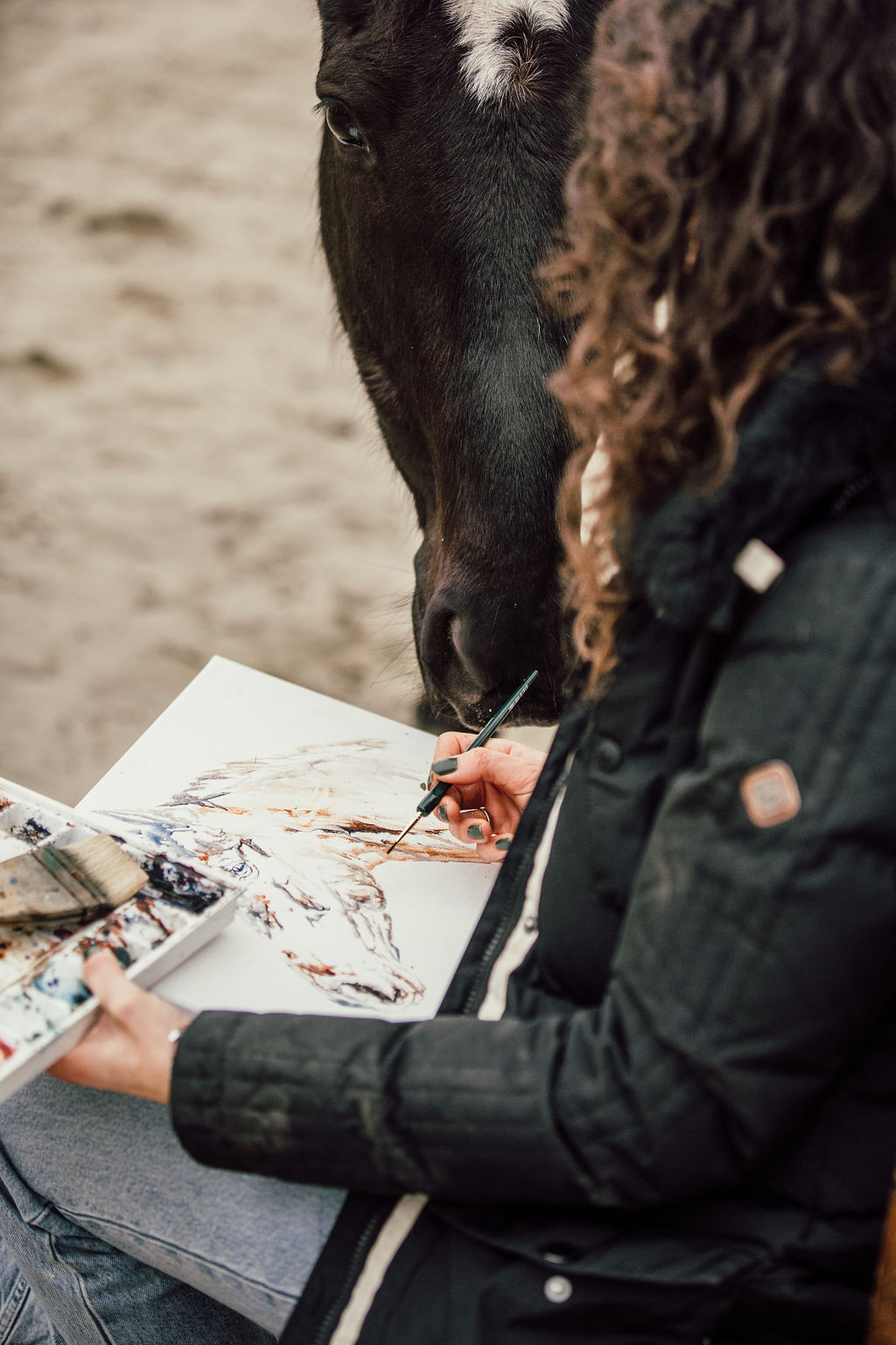 Marjo painting on a board while a black horse leans in close beside her