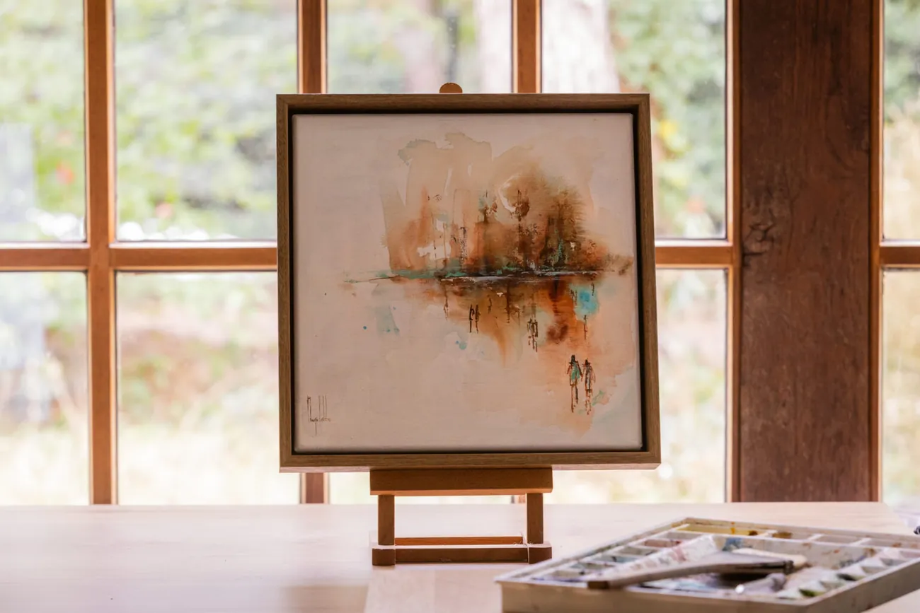 The framed painting on a small easel in the studio, in front of a window, beside an open watercolour box