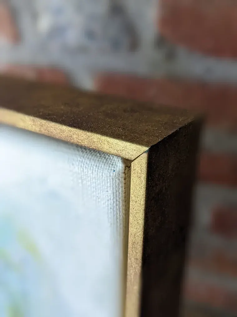 The corner of Aurora's wooden floating frame in its gold brass finish, the canvas edge visible inside it
