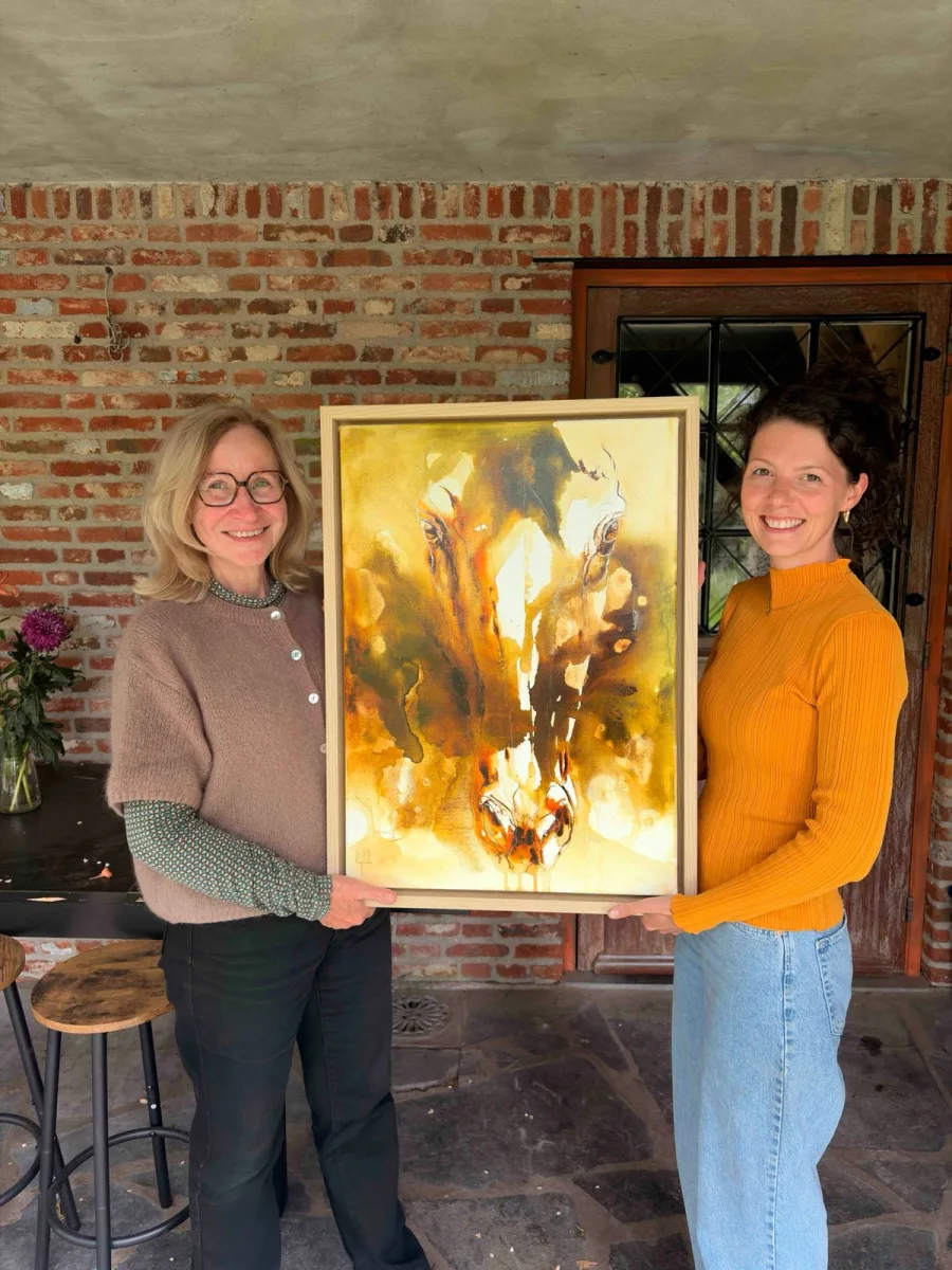 Marjo Lebbe en de koper houden een verkocht ingekaderd schilderij uit de reeks Coalescence vast tegen een bakstenen muur
