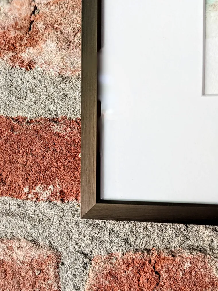 The corner of the brushed aluminium frame and its white mat against a brick wall