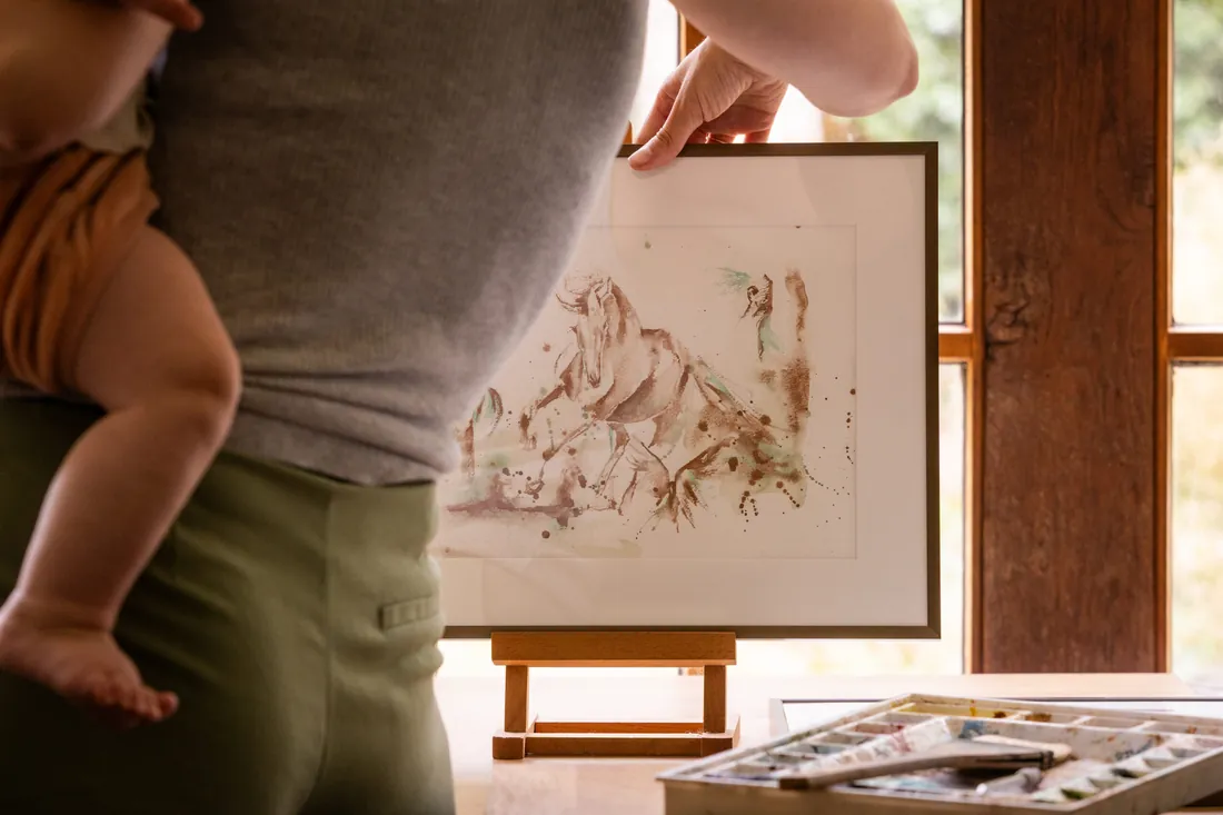Marjo Lebbe holding the framed Equus et Avis by the studio window, her watercolour palette in the foreground