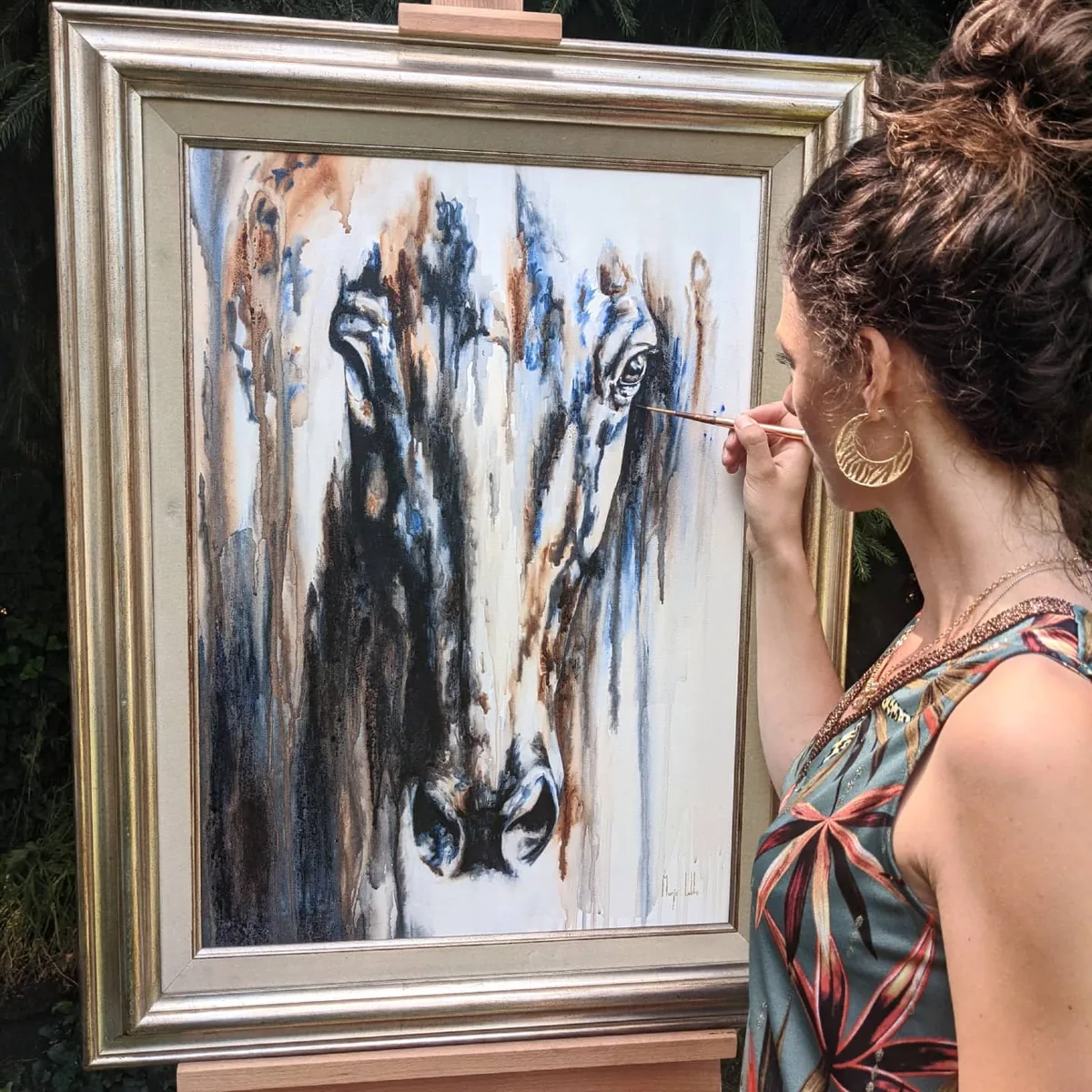 Marjo aan het werk aan het schilderij: het ingelijste doek staat buiten op een ezel tegen het groen, haar penseel bij het open oog van het paard