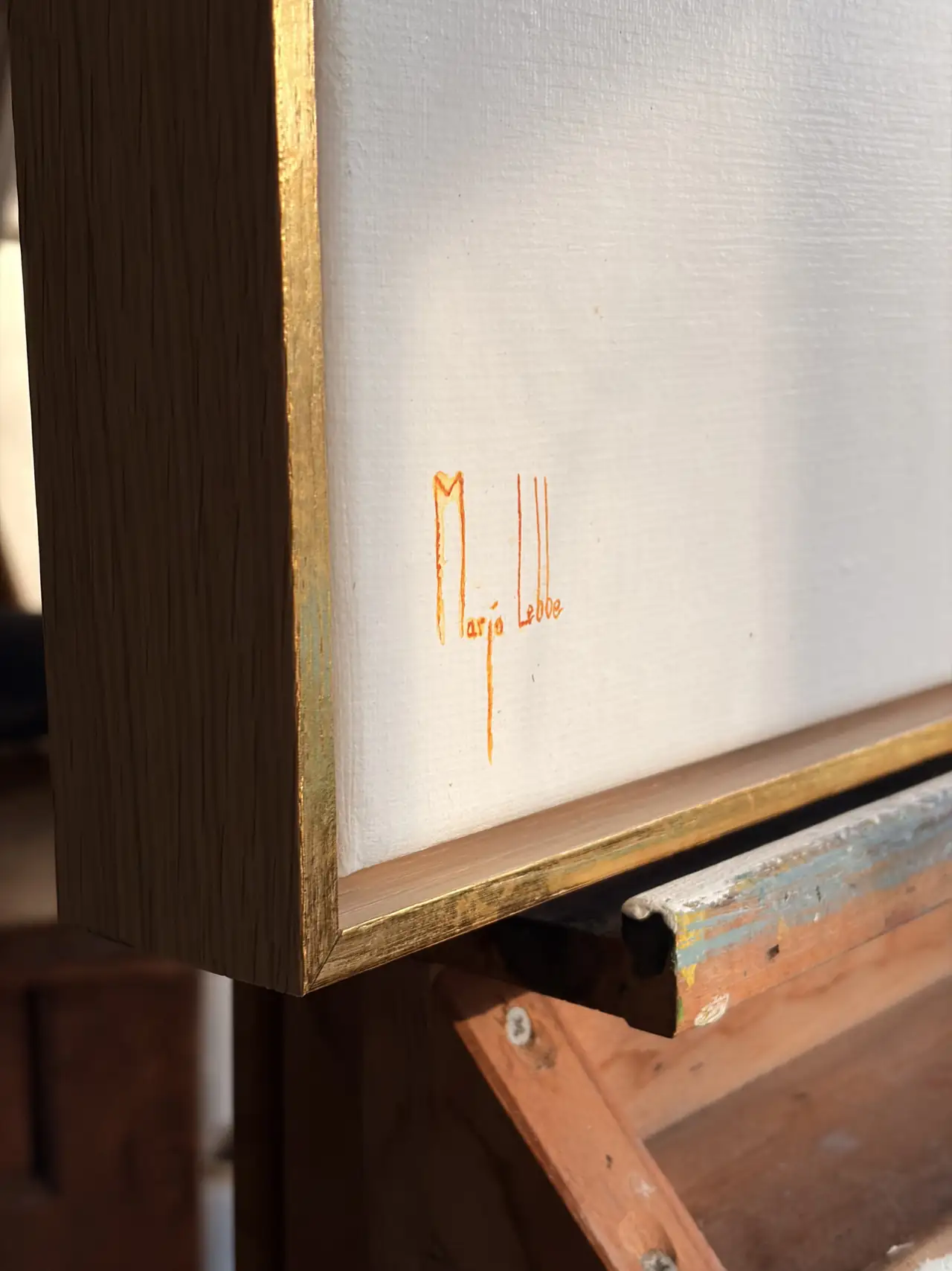 De handtekening van Marjo Lebbe in oranje aquarel, laag op het witte linnen, de gouden lijstrand vangt het licht