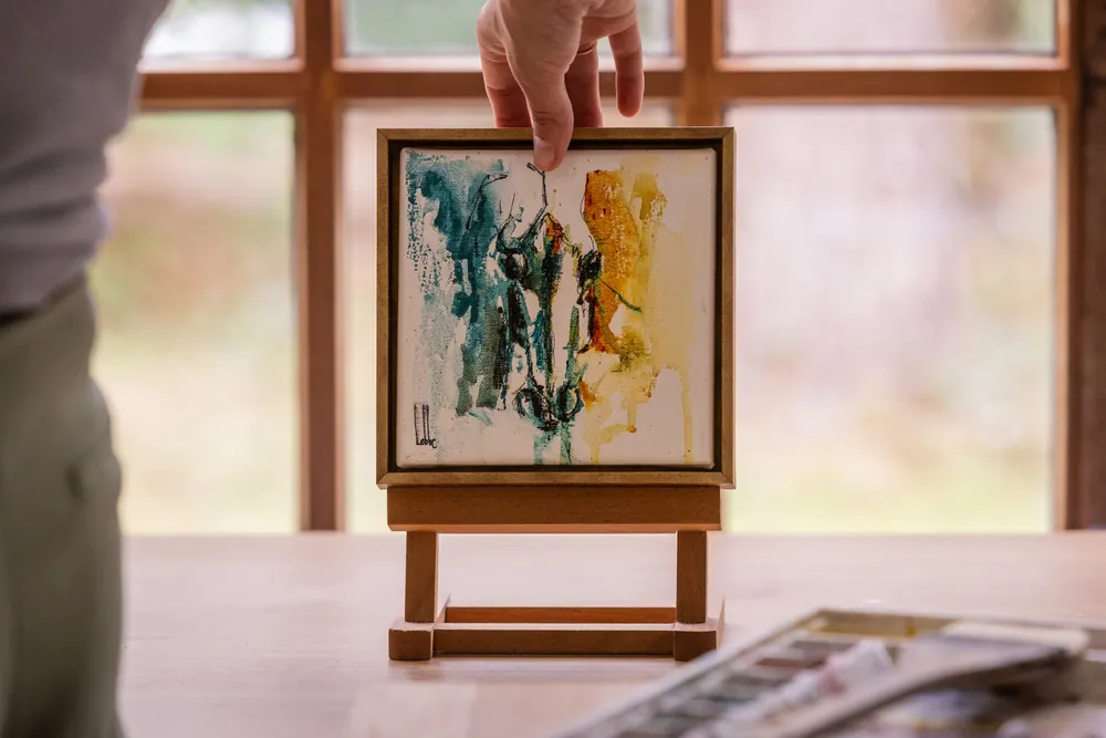 Een hand die het blauw- en okerkleurige Small things aquarel van een ezeltje tilt in het atelier